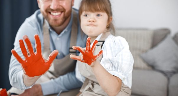 Little girl with Down syndrome wearing white shirt and beige apron. Girl's and her father's hands painted with red paint. Father and his daughter showing their hands.